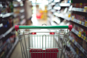 Carrinho de compras vazio em um corredor de supermercado, com prateleiras repletas de produtos desfocados ao fundo.
