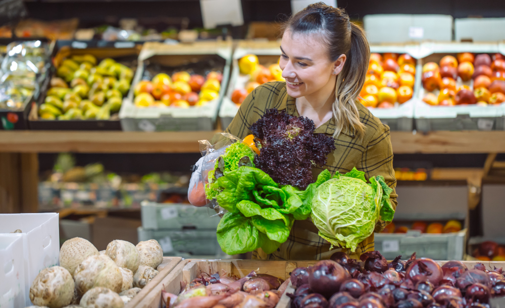 Mulher sorridente segurando diversas folhas verdes frescas em uma feira ou seção de hortifrúti de supermercado.