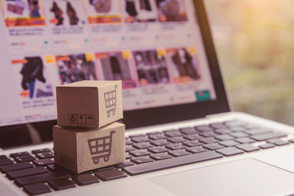 Dois mini pacotes de papelão com ícones de carrinho de compras sobre o teclado de um notebook exibindo uma loja virtual na tela.