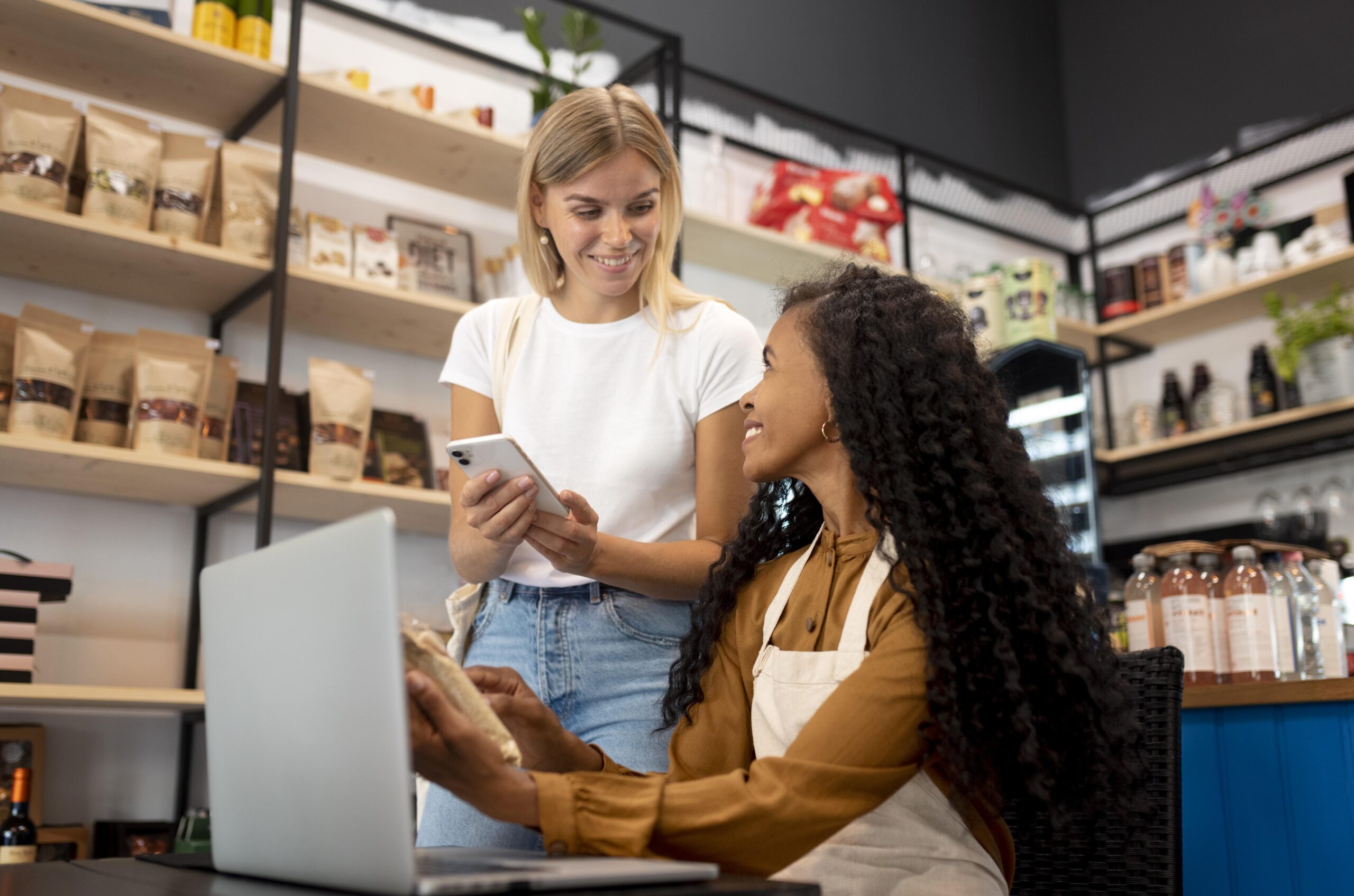 Duas mulheres sorrindo em uma loja; uma delas está em pé com um celular na mão e a outra sentada com um notebook e um pacote de produto, interagindo de forma amigável.