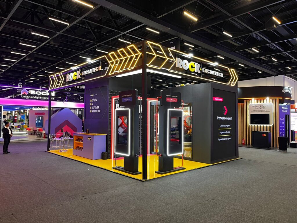Estande iluminado da Rock EncanTech em feira de tecnologia, com painéis digitais, setas de LED e design moderno, cercado por outros estandes.