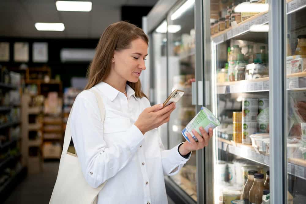 Mulher escaneando produto com o celular em frente a geladeira de supermercado.