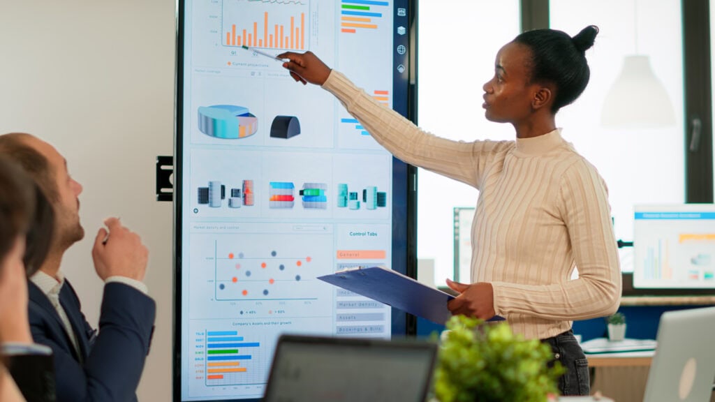 Mulher em reunião corporativa apresentando dados e gráficos em uma tela digital para colegas, ilustrando a importância de decisões data-driven no varejo.