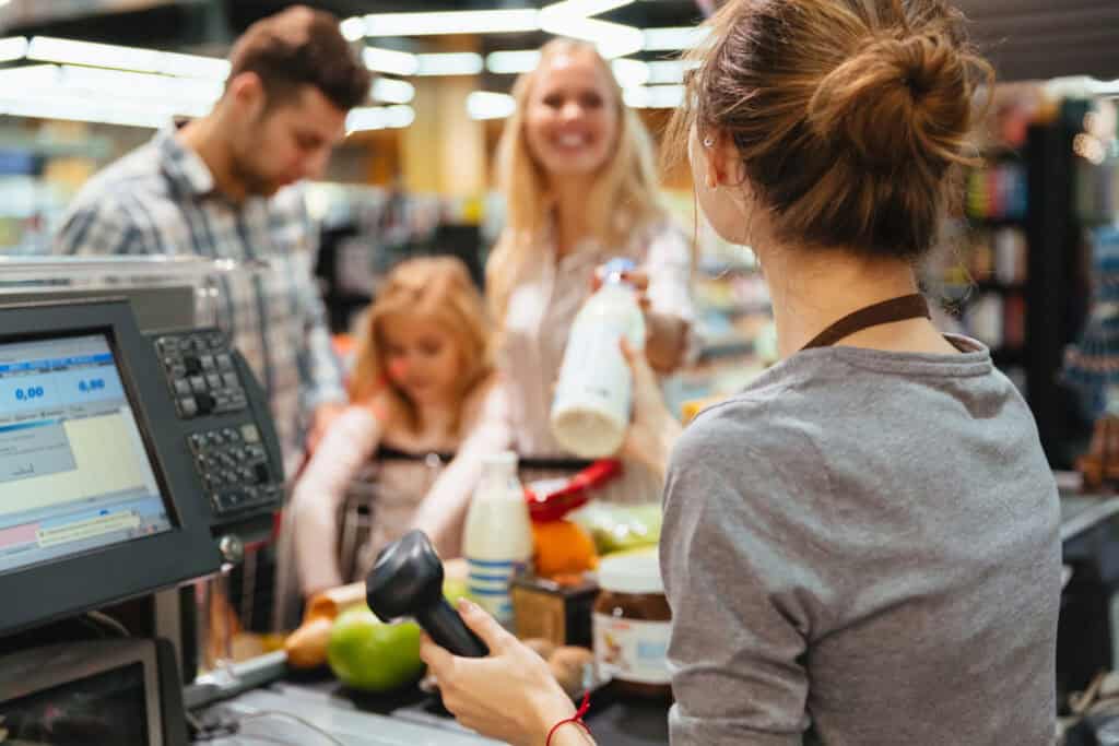 Caixa de supermercado atendendo uma família sorridente no caixa. A funcionária segura o leitor de código de barras enquanto uma cliente entrega uma garrafa de leite, com outras compras visíveis na esteira.