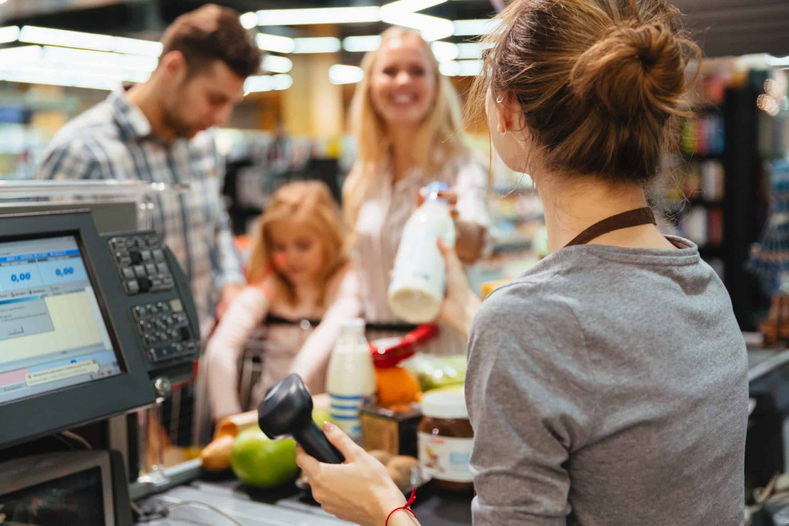 Caixa de supermercado atendendo uma família sorridente no caixa. A funcionária segura o leitor de código de barras enquanto uma cliente entrega uma garrafa de leite, com outras compras visíveis na esteira.