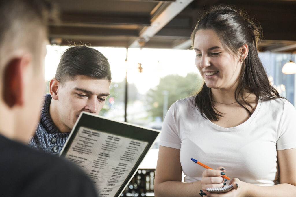 Garçonete sorridente anota o pedido de clientes em um restaurante enquanto um deles lê o cardápio.