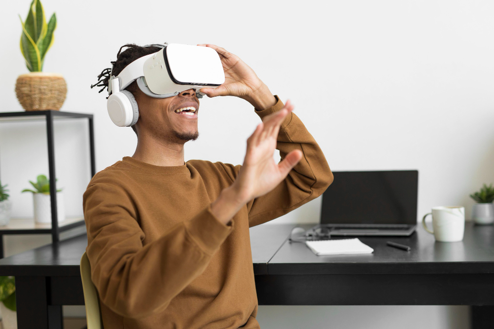 Pessoa sorrindo e usando óculos de realidade virtual enquanto interage com as mãos em um escritório com mesa, laptop e caderno ao fundo.