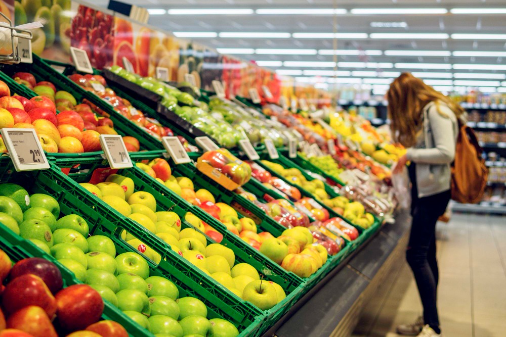 Seção de hortifrúti de supermercado com maçãs organizadas em bandejas, enquanto uma mulher escolhe frutas ao lado.