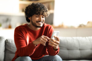 Pessoa sentada em um sofá, sorrindo enquanto usa um smartphone, em um ambiente doméstico iluminado.