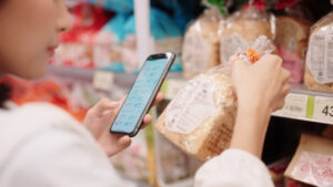 Uma mulher em um supermercado, segurando um smartphone em uma mão e um pacote de pão na outra, olhando a embalagem e a tela do telefone.