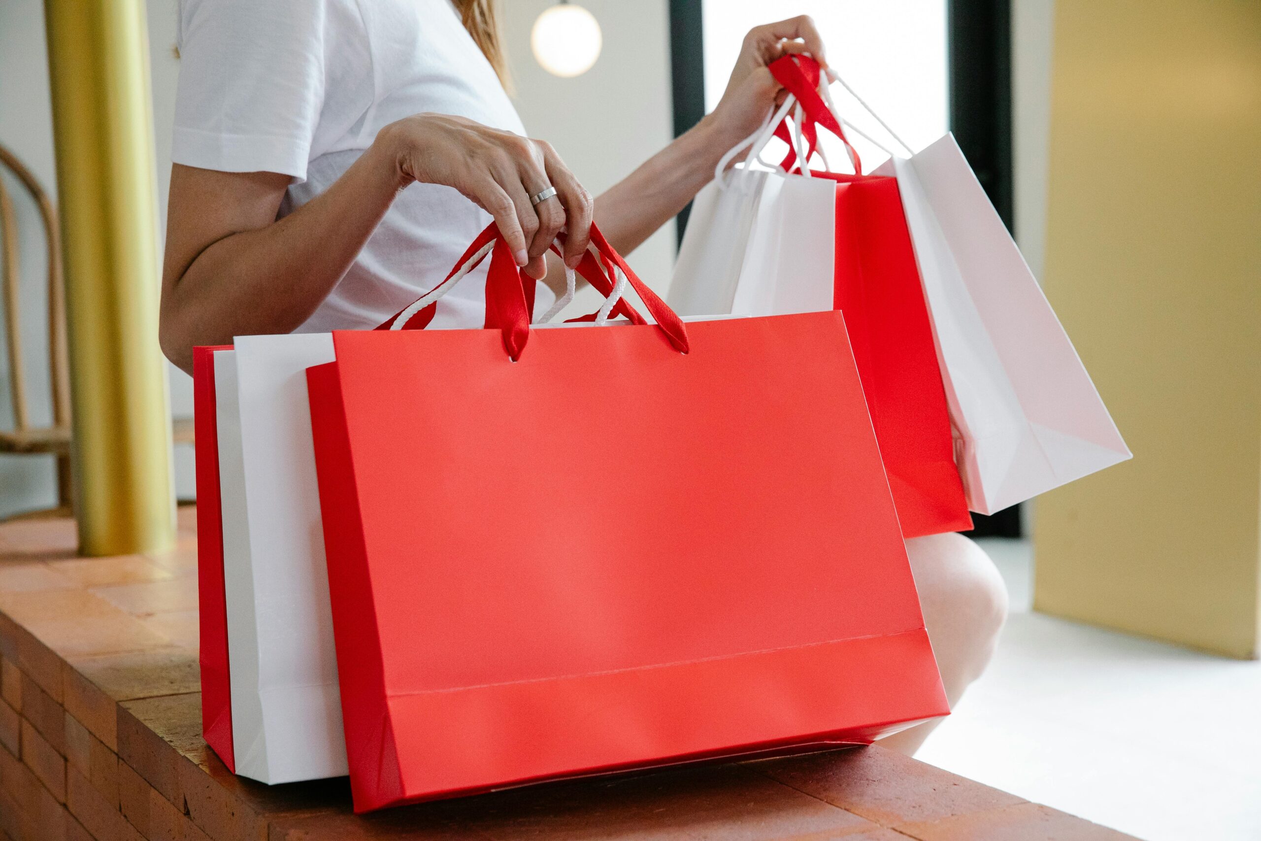 Pessoa segurando várias sacolas de compras vermelhas e brancas em ambiente interno, representando consumo e varejo.