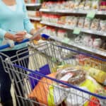 Pessoa fazendo lista de compras em supermercado ao lado de carrinho cheio de produtos em corredor de alimentos.