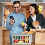 Casal organiza compras de supermercado enquanto consulta o celular na cozinha de casa.