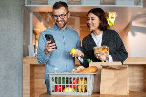 Casal organiza compras de supermercado enquanto consulta o celular na cozinha de casa.