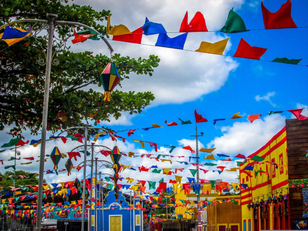 Decoração tradicional de festas juninas com bandeirinhas coloridas, balões decorativos e fachadas típicas em uma rua enfeitada, representando a sazonalidade que movimenta o varejo alimentar e aumenta a procura por alimentos tradicionais.