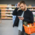 Cliente homem em um corredor de supermercado segura uma cesta de compras laranja e olha para um bloco de notas com expressão de dúvida, coçando a cabeça. Ao fundo, prateleiras com produtos.