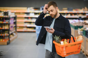 Cliente homem em um corredor de supermercado segura uma cesta de compras laranja e olha para um bloco de notas com expressão de dúvida, coçando a cabeça. Ao fundo, prateleiras com produtos.