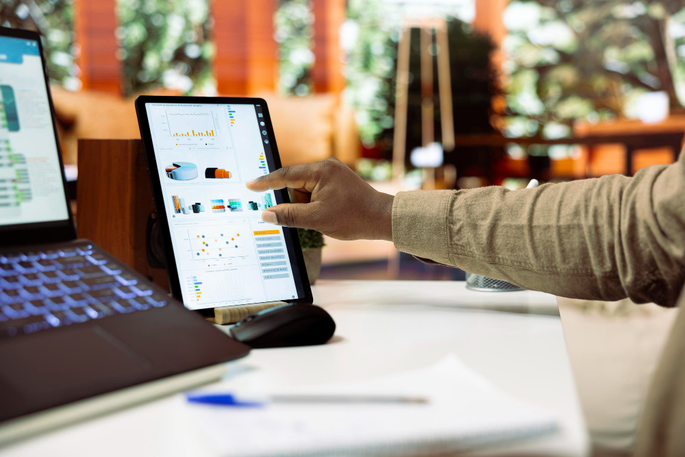 Mão de profissional interagindo com tablet que exibe gráficos e dashboard de análise de dados sobre mesa de trabalho com laptop.