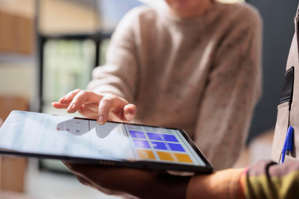 Fotografia em plano fechado de duas pessoas a analisar dados num tablet. Uma pessoa segura o equipamento enquanto a outra aponta o dedo para um painel com blocos coloridos de informação no ecrã.
