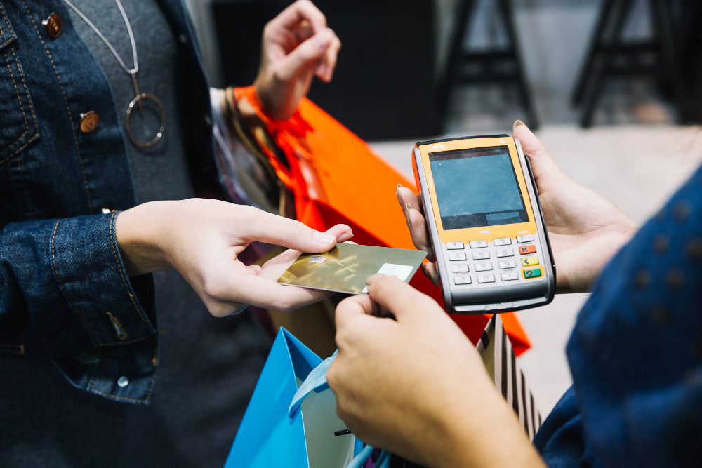Cliente entrega um cartão de crédito dourado ao operador de caixa de um supermercado para realizar o pagamento, com compras e gôndolas ao fundo.