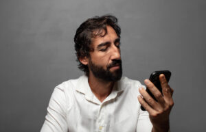 Homem de barba e camisa branca olhando para a tela de um celular.