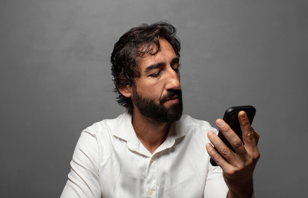 Homem de barba e camisa branca olhando para a tela de um celular.