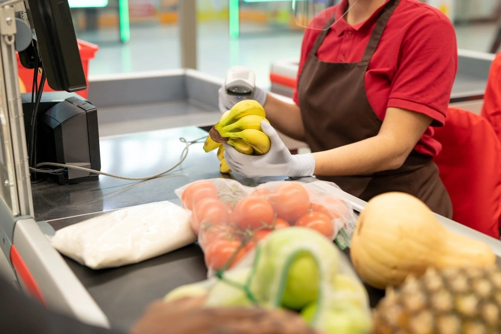 Operadora de caixa de supermercado, vestindo uniforme vermelho, avental marrom e luvas brancas, segura bananas e um leitor de código de barras durante o atendimento, com frutas e verduras sobre a esteira.