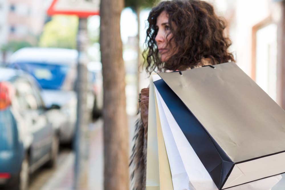 A imagem mostra uma mulher com cabelos escuros e encaracolados a caminhar num passeio urbano, vista de perfil e virada parcialmente de costas. Em primeiro plano, destaca-se um conjunto de sacos de compras de papel — de cores como preto, branco e bege — que ela carrega ao ombro. Ela apresenta uma expressão séria e pensativa enquanto olha para o lado. O fundo da cena, composto por carros estacionados, o tronco de uma árvore e a arquitetura de uma rua da cidade, encontra-se desfocado sob uma iluminação natural e suave.